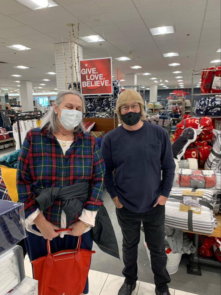 Ryan and Lynn's mom at Macy's with an industrial sheet-buying trolley, a 'Give. Love. Believe.' sign above them